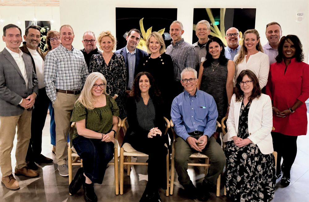 A group of about twenty adults gathered indoors for a posed group photo. Four people are seated at the front, with the rest standing behind them. Everyone is smiling, and the setting is an event space with artwork on the walls.