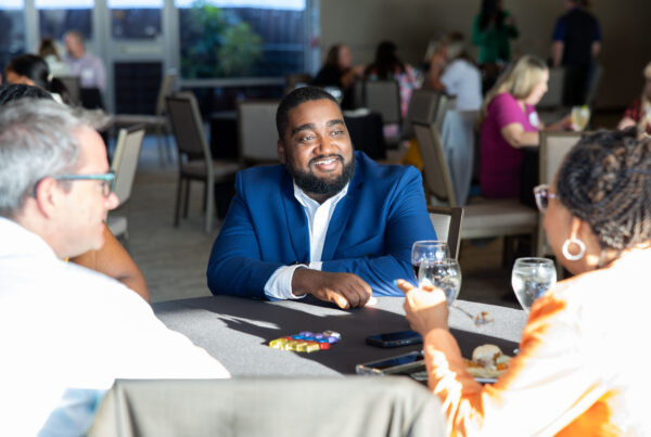 Three adults sit at a table talking, with a smiling man in a blue suit at the center.