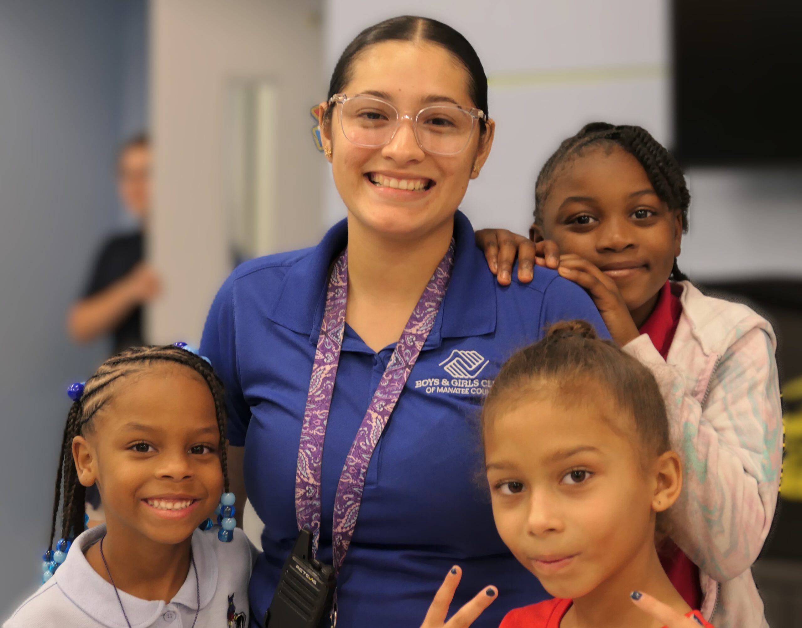 A smiling adult wearing glasses and a blue Boys & Girls Club polo stands with three young girls gathered around her, all posing closely together indoors in what appears to be a classroom or community center.