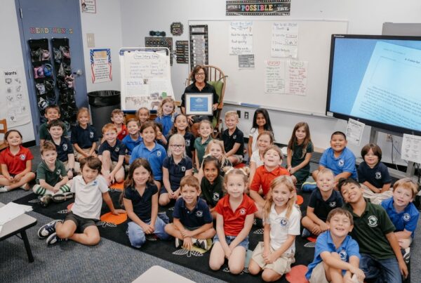 Teacher holding a Ripple Effect Teacher Award certificate sits in a classroom with a large group of smiling elementary students gathered around her.